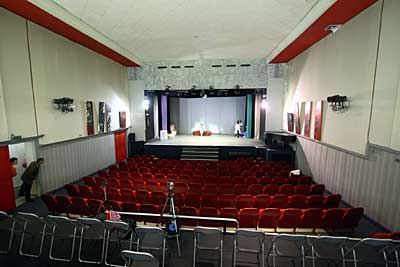 Theater Maestro Bühne, Standpunkt der Aufnahme ist der Zuschauerraum, zu sehen ist eine Kastenbühne mit Stiegenaufgang, den Zuschauerraum zieren rot gepolsterte Sessel.
